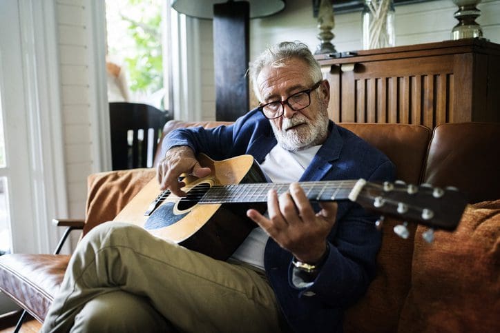 Elderly man playing acoustic guitar indoors with a relaxed expression.